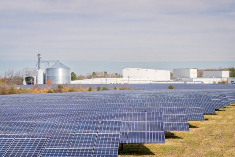 Planta fotovoltaica | Autoconsumo fotovoltaico a gran escala 14 A large solar panel field with warehouses and silos in the background under a clear sky.